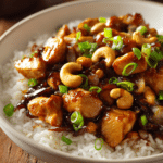 Close-up of Authentic Cashew Chicken stir-fry with golden brown chicken cubes, whole raw cashews, glossy dark soy-based sauce, and bright green sliced onions in a shallow white bowl on a rustic wooden surface