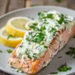 Close-up of baked salmon fillet topped with creamy garlic and herb Boursin cheese, garnished with fresh parsley and a thin lemon wedge on a white plate with warm natural lighting