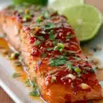 Close-up of baked salmon fillet with sticky sweet chili garlic glaze, garnished with chopped cilantro and lime wedges on a white rectangular plate