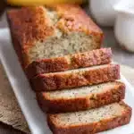 Close-up of rustic, golden brown classic banana bread with visible banana specks, sliced and arranged on a white plate, showcasing moist, tender crumb and slightly crisp crust in warm natural light