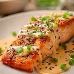 Pan-seared salmon fillet with crispy exterior topped with creamy spicy bang bang sauce, garnished with chopped green onions and black and white sesame seeds on a white plate
