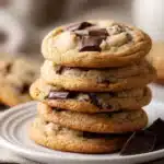 Close-up of soft and chewy classic chocolate chip cookies stacked on a white ceramic plate, showing melted semi-sweet chocolate chips and golden brown edges under warm natural light