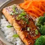 Close-up of pan-seared salmon fillet with crispy golden brown skin on white jasmine rice, garnished with green onions and sesame seeds, alongside steamed broccoli and carrot ribbons with soy-ginger glaze drizzle