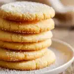 Close-up of softly baked sugar cookies stacked on a white ceramic plate with powdered sugar dusting, showcasing soft and chewy texture and golden edges in warm natural light.