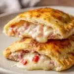 Close-up image of two homemade hot pockets with golden flaky crust and melted mozzarella cheese, cooked diced ham, diced bell peppers, and onions on a white round ceramic plate