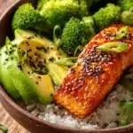 Close-up of honey glazed salmon fillet with crispy exterior on a bed of jasmine rice, steamed broccoli florets, sliced avocado, sesame seeds, and sliced green onions on a rustic wooden surface with warm natural light.
