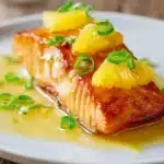 Close-up of pan-seared and oven-baked salmon fillet with crispy golden brown crust, glazed with bright pineapple sauce, garnished with thin pineapple slices and chopped green onions on a white plate.