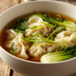 Close-up of potsticker soup served in a white bowl with soft dumplings, bright green bok choy and scallions in a clear soy-ginger broth, garnished with sesame oil drizzle