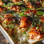 Close-up of spicy salmon sushi bake in a rectangular baking dish showing layers of cooked sushi rice, salmon mixed with spicy mayo, garnished with green onions and furikake seasoning on rustic surface