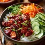 Close-up of a layered chicken bowl with tender chicken thighs coated in glossy red gochujang sauce, served over white rice, with julienned carrots, sliced cucumbers, green onions, and sesame seeds on a rustic wooden surface.