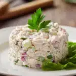 Close-up of classic tuna salad with creamy texture, finely chopped celery and red onion, garnished with a bright green parsley sprig, served on a white plate with warm natural lighting.