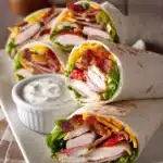 Close-up of turkey ranch club wraps cut diagonally on a white plate, showing layers of sliced turkey breast, crispy bacon, leafy romaine lettuce, sliced tomatoes, shredded cheddar cheese, and ranch dressing with warm natural light