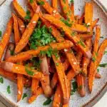 Air fryer garlic sweet potato fries - the image shows a plate of cooked sweet potato fries. the fries are orange in color and have a crispy texture. they are garnished with chopped parsley and sesame seeds. the plate is white with a speckled pattern and is sitting on a wooden table. the background is blurred, but it appears to be a kitchen countertop.