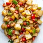 Air fryer potato hash - the image shows a pile of diced potatoes on a white marble countertop. the potatoes are golden brown and appear to be seasoned with herbs and spices. there are also small pieces of red and green bell peppers scattered throughout the dish. the dish is garnished with chopped parsley. the overall appearance of the dish is colorful and appetizing.