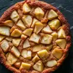 Apple cake one bowl - the image is a close-up of a round, golden-brown tart with a crumbly texture. the tart is placed on a dark, textured surface, possibly a table or countertop. on top of the tart, there are small cubes of sliced apples arranged in a grid-like pattern. the apples appear to be caramelized and have a golden brown color, indicating that they have been freshly baked. the crust is golden brown and has a slightly crispy texture.