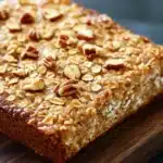 Banana bread baked oatmeal - the image shows a rectangular piece of bread on a wooden cutting board. the bread appears to be freshly baked and has a golden brown crust on top. it is topped with chopped nuts, giving it a crunchy texture. the nuts are scattered across the top of the bread, adding a pop of color to the dish. the cutting board is placed on a dark grey countertop.