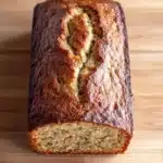 Banana cake loaf photography - the image shows a loaf of bread on a wooden cutting board. the bread appears to be freshly baked and has a golden brown crust on top. the inside of the bread is visible, with a crumbly texture and a soft, fluffy interior. the loaf is cut in half, revealing the soft, golden-brown interior of the loaf. the cutting board has a light-colored wood grain pattern. the background is blurred, making the bread the focal point of the image.
