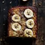 Banana cake slice plating - the image is a close-up of a square-shaped cake on a wooden surface. the cake appears to be freshly baked and has a golden brown crust on top. on top of the cake, there are several sliced bananas arranged in a circular pattern. the bananas are cut into thin slices and are arranged in an overlapping manner. there is a single banana peel on the right side of the image, which is partially visible in the top right corner. the background is dark and out of focus, making the cake stand out.