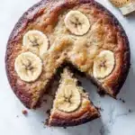 Banana cake toddler - the image shows a round cake with a slice taken out of it. the cake is golden brown in color and has a crumbly texture. on top of the cake, there are several sliced bananas arranged in a circular pattern. the bananas are cut in half, revealing the soft, fluffy interior. there are also a few whole bananas scattered around the cake. the background is a white marble countertop.