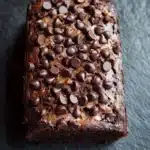 Banana chocolate cake - the image is a close-up of a square-shaped chocolate cake. the cake appears to be freshly baked and has a golden brown color. it is topped with small chocolate chips scattered across the top, giving it a shiny and shiny appearance. the background is a dark grey textured surface, making the cake stand out. the overall mood of the image is dark and decadent.