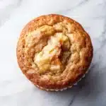 Banana muffin gluten free - the image is a close-up of a muffin on a white marble surface. the muffin appears to be freshly baked and has a golden brown color. it has a crumbly texture on top, with a swirl of butter on top. the butter is a light yellow color and looks soft and fluffy. the background is blurred, making the muffin the focal point of the image.