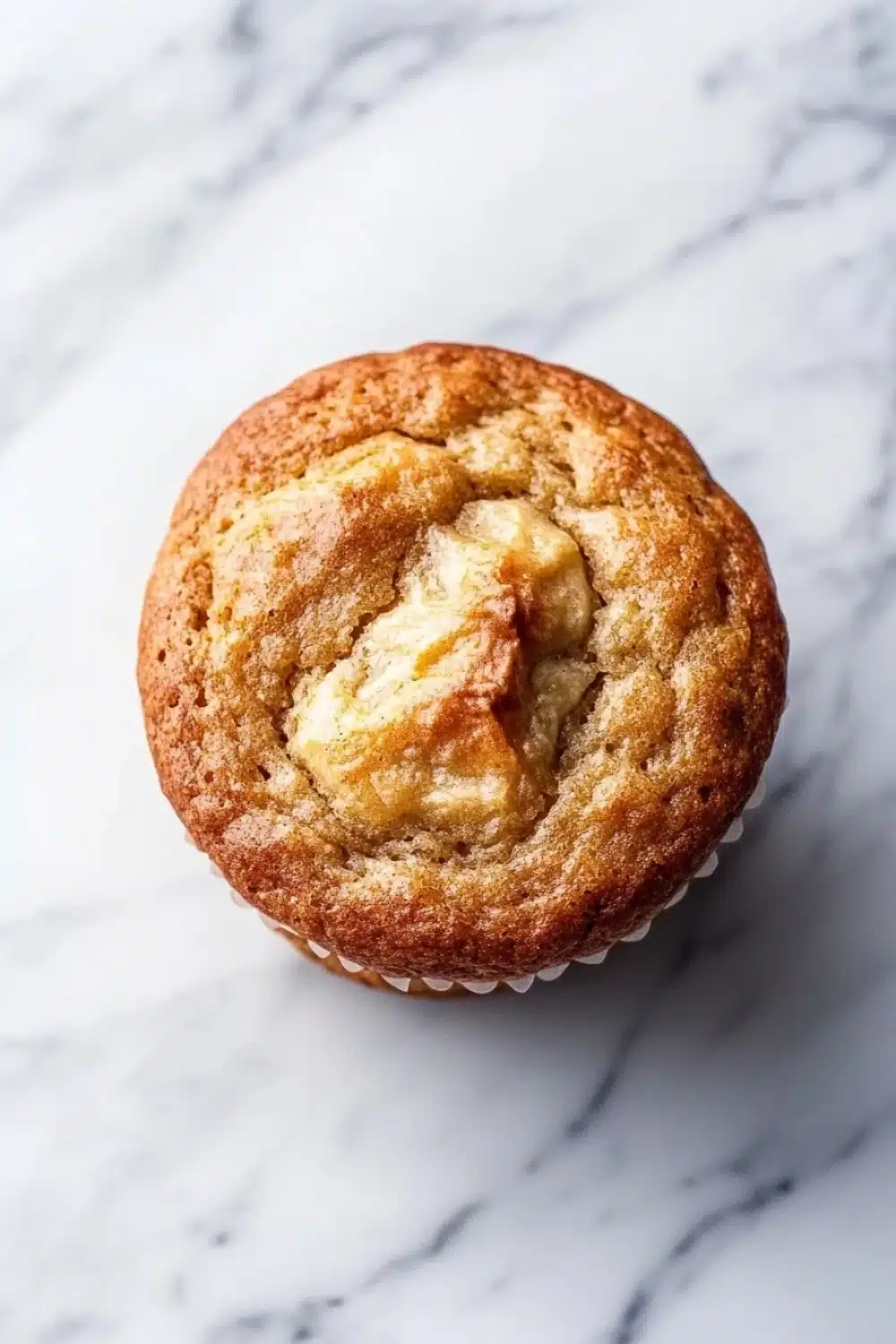 Banana muffin gluten free - the image is a close-up of a muffin on a white marble surface. the muffin appears to be freshly baked and has a golden brown color. it has a crumbly texture on top, with a swirl of butter on top. the butter is a light yellow color and looks soft and fluffy. the background is blurred, making the muffin the focal point of the image.