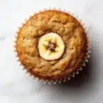 Banana muffin no added sugar - the image is a close-up of a freshly baked muffin on a white marble surface. the muffin is golden brown in color and has a crumbly texture. on top of the muffin, there is a single banana cut in half, revealing the soft, fluffy interior. the banana is covered in a light dusting of powdered sugar, giving it a shiny appearance. the background is blurred, making the muffins the focal point of the image.