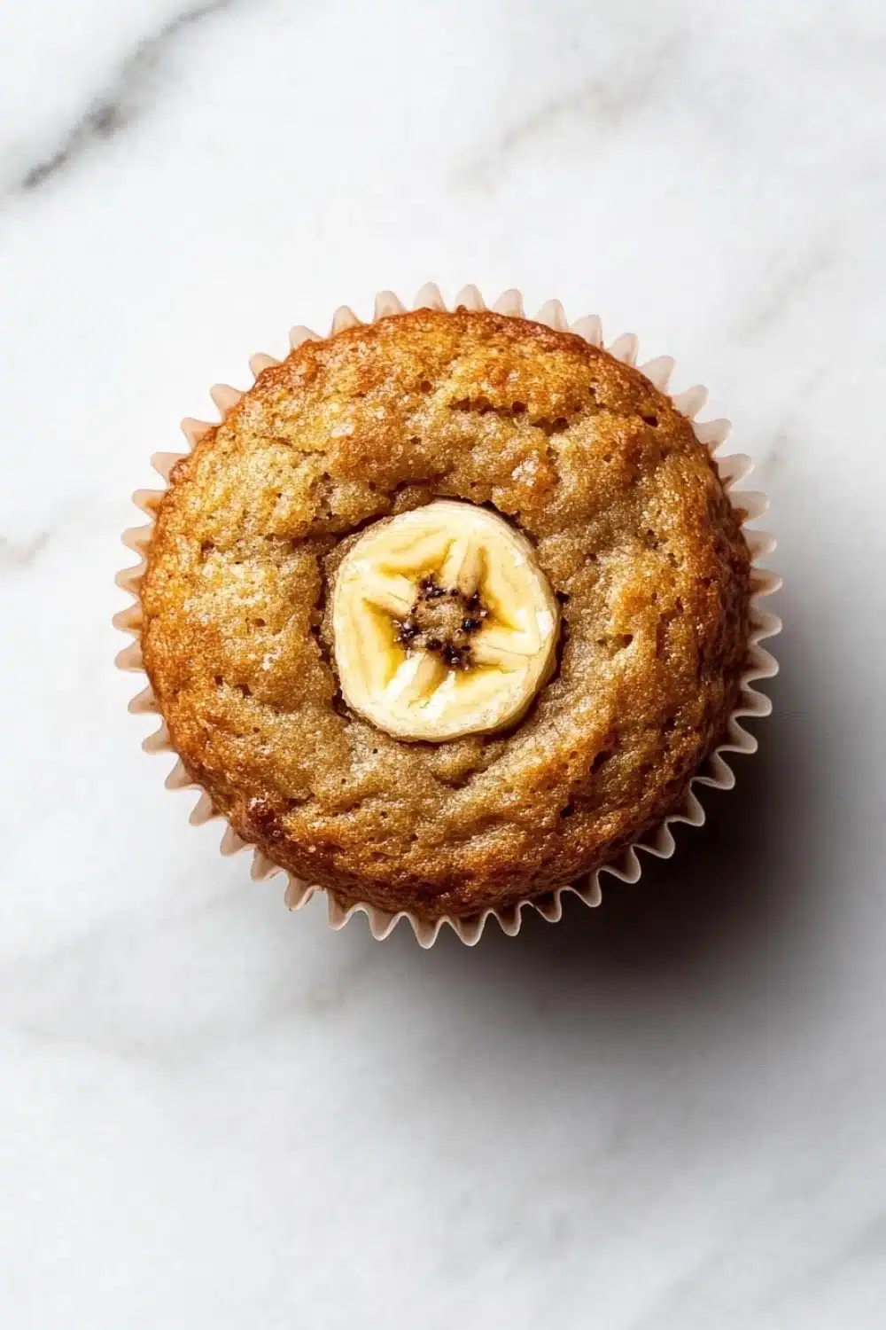 Banana muffin no added sugar - the image is a close-up of a freshly baked muffin on a white marble surface. the muffin is golden brown in color and has a crumbly texture. on top of the muffin, there is a single banana cut in half, revealing the soft, fluffy interior. the banana is covered in a light dusting of powdered sugar, giving it a shiny appearance. the background is blurred, making the muffins the focal point of the image.