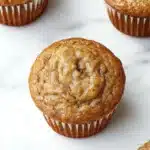 Banana muffin protein - the image shows a group of freshly baked muffins on a white marble surface. there are six muffins in total, arranged in a row. the muffins are golden brown in color and have a crumbly texture. they are topped with a generous amount of cinnamon sugar, giving them a warm and inviting appearance. the background is blurred, making the muffins the focal point of the image.