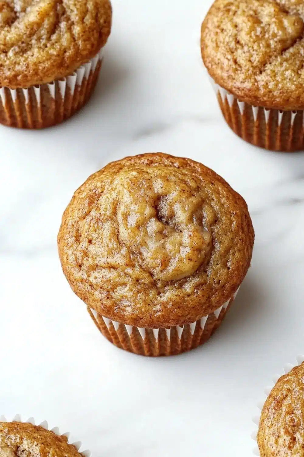 Banana muffin protein - the image shows a group of freshly baked muffins on a white marble surface. there are six muffins in total, arranged in a row. the muffins are golden brown in color and have a crumbly texture. they are topped with a generous amount of cinnamon sugar, giving them a warm and inviting appearance. the background is blurred, making the muffins the focal point of the image.
