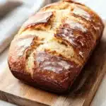 Banana muffin rustic bread - the image shows a loaf of bread on a wooden cutting board. the bread appears to be freshly baked, with a golden brown crust and a soft, fluffy interior. the loaf is sitting on a white marble countertop, with the cutting board resting on top of it. a white cloth napkin is visible in the background, suggesting that the bread is ready to be served. the overall mood of the image is rustic and cozy.