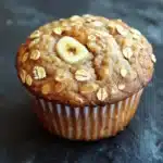 Banana oat yogurt muffin - the image is of a freshly baked muffin on a dark grey textured surface. the muffin is golden brown in color and has a crumbly texture. on top of the muffin, there is a single banana cut in half, revealing the soft, fluffy interior. the banana is cut in a way that it looks like it has been freshly baked and is placed on top of a pile of oats. the oats are scattered around the banana, adding a crunchy texture to the dish. the background is blurred, making the muffins the focal point of the image.