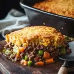 Beef casserole with cornbread topping - the image shows a slice of shepherd's pie on a wooden cutting board. the pie has a golden brown crust on top and is filled with ground beef, peas, carrots, and green onions. a spoon is resting on the cutting board next to the pie. in the background, there is a black casserole dish with a white cloth napkin. the background is dark and out of focus.
