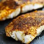 Blackened tilapia in air fryer - the image shows two pieces of fish fillets on a black slate plate. the fillets are golden brown in color and appear to be seasoned with spices. the seasoning is sprinkled on top of the fillets, giving them a crispy texture. the background is blurred, but it appears to be a kitchen countertop. the overall mood of the image is warm and inviting.