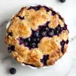 Blueberry muffin mix cobbler - the image is of a freshly baked blueberry pie in a white ceramic pie dish. the pie has a golden brown crust on top and is filled with fresh blueberries. the blueberries are arranged in a circular pattern around the edges of the pie. the dish is sitting on a white marble countertop with a few scattered blueberries around it. the overall aesthetic of the image is fresh and appetizing.