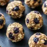 Blueberry muffin protein balls - the image shows a tray of freshly baked oatmeal balls. the balls are round and golden brown in color, with visible oats and blueberries scattered throughout. they are arranged in a scattered manner on a black surface, with some overlapping each other. the background is blurred, but it appears to be a kitchen countertop.