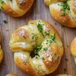 Bread knots - the image shows a wooden cutting board with several freshly baked pretzels arranged in a circular pattern. the pretzes are golden brown in color and have a soft, fluffy texture. they are topped with a generous amount of melted cheese and a sprig of fresh parsley. the cheese appears to be melted and bubbly, and the parsley adds a pop of green color to the dish. the background is blurred, but it seems to be a kitchen countertop.