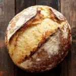 Bread with self rising flour - the image is a close-up of a freshly baked loaf of bread on a wooden surface. the bread appears to be freshly baked, with a golden brown crust and a soft, fluffy interior. the edges of the bread are slightly frayed, indicating that it has been freshly baked. the wood has a weathered and aged appearance, with visible grain and knots. the background is blurred, making the bread the focal point of the image.