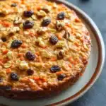 Carrot cake oatmeal bake - the image is a close-up of a round cake on a white plate with a brown rim. the cake appears to be a carrot cake with a golden-brown crust and is topped with chopped nuts and raisins. the nuts are scattered around the plate, and there are a few scattered nuts on the table in the background. the plate is sitting on a dark grey surface.