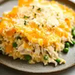 Cheesy chicken casserole - the image shows a plate of food with a slice taken out of it. the dish appears to be a casserole or lasagna, with a golden brown crust on top. the top of the dish is covered in a layer of melted cheese, which is a bright orange color. the cheese is sprinkled with chopped herbs, giving it a crunchy texture. the plate is brown and has a speckled pattern around the edges. there are also a few small green peas scattered around the plate. the background is a white marble countertop.