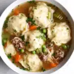Chicken and beef dumplings crockpot freezer meal - the image is a close-up of a bowl of soup. the soup appears to be a beef noodle soup with dumplings, carrots, peas, and meat. the bowl is white and is sitting on a white marble countertop. the broth is a light brown color and there are small chunks of meat visible in the soup. there are also some green herbs sprinkled on top of the soup, adding a pop of color to the dish.