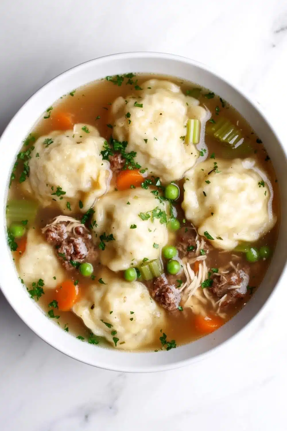 Chicken and beef dumplings crockpot freezer meal - the image is a close-up of a bowl of soup. the soup appears to be a beef noodle soup with dumplings, carrots, peas, and meat. the bowl is white and is sitting on a white marble countertop. the broth is a light brown color and there are small chunks of meat visible in the soup. there are also some green herbs sprinkled on top of the soup, adding a pop of color to the dish.