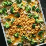 Chicken and broccoli casserole - the image shows a white rectangular baking dish filled with a casserole dish. the dish appears to be made with broccoli, chicken, and cheese. the broccoli is cut into small pieces and is arranged in a circular pattern on top of the chicken. the cheese is melted and bubbly, and there are crumbled breadcrumbs scattered throughout the dish. there are a few more broccoli florets scattered around the dish, adding a pop of green color to the image. the background is a dark wooden table, and the overall color scheme is warm and inviting.