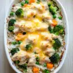 Chicken and rice with veggies casserole oven - the image shows a white ceramic baking dish filled with a casserole dish. the dish is filled with rice, broccoli, peas, carrots, and chunks of chicken. the chicken is covered in a layer of melted cheese, which is golden brown and bubbly. the broccoli is bright green and the peas are small and round. the rice is white and fluffy, and the dish is sitting on a wooden table. the background is blurred, but it appears to be a kitchen countertop.