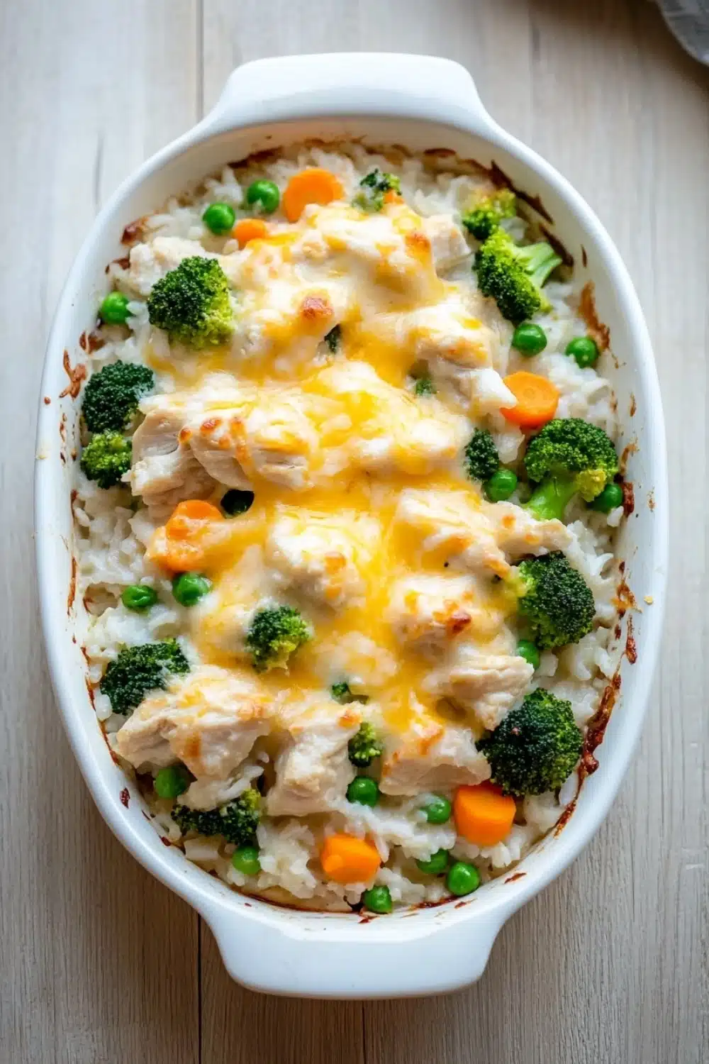 Chicken and rice with veggies casserole oven - the image shows a white ceramic baking dish filled with a casserole dish. the dish is filled with rice, broccoli, peas, carrots, and chunks of chicken. the chicken is covered in a layer of melted cheese, which is golden brown and bubbly. the broccoli is bright green and the peas are small and round. the rice is white and fluffy, and the dish is sitting on a wooden table. the background is blurred, but it appears to be a kitchen countertop.