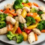 Chicken and vegetables freezer meal - the image is a close-up of a plate of stir-fry with chicken and vegetables. the plate is round and has a gray rim. the chicken is cut into small pieces and is mixed with various vegetables such as broccoli, red and yellow bell peppers, and orange carrots. the vegetables are cut into thin strips and are arranged in a colorful and appetizing manner. the dish appears to be freshly cooked and ready to eat.