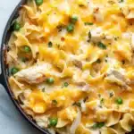 Chicken noodle casserole skillet - the image shows a black cast iron skillet filled with a creamy, yellow-orange dish. the dish appears to be a macaroni and cheese casserole, with chunks of chicken and peas scattered throughout. the cheese is melted and bubbly, and there are small green herbs sprinkled on top. the skillet is sitting on a white marble countertop.