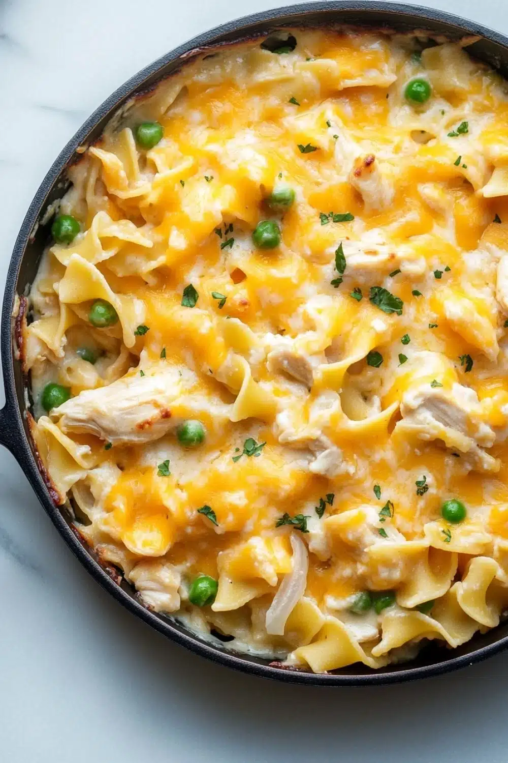 Chicken noodle casserole skillet - the image shows a black cast iron skillet filled with a creamy, yellow-orange dish. the dish appears to be a macaroni and cheese casserole, with chunks of chicken and peas scattered throughout. the cheese is melted and bubbly, and there are small green herbs sprinkled on top. the skillet is sitting on a white marble countertop.