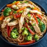 Chicken noodles low calorie - the image is a close-up of a dish in a black bowl. the dish appears to be a stir-fry with noodles, vegetables, and chicken. the noodles are light brown and appear to be cooked. the vegetables include broccoli, red bell peppers, and sliced carrots. the chicken is cut into small pieces and is scattered throughout the dish. the colors of the vegetables are vibrant and the dish looks fresh and appetizing. the bowl is sitting on a dark, textured surface.