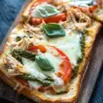 Chicken pesto french bread pizza - the image shows a wooden cutting board with three slices of bread on it. the bread appears to be toasted and has a golden brown crust. on top of the bread, there is a generous amount of melted cheese, which is topped with shredded chicken, sliced tomatoes, and fresh basil leaves. the tomatoes are bright red and appear to be ripe and juicy. the basil leaves are a vibrant green color and are scattered throughout the dish. the background is dark, making the bread and cheese stand out.